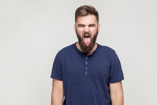 Crazy and fun concept. Bearded young adult man showing tongue at camera and have a happiness look. Studio shot, isolated on gray background