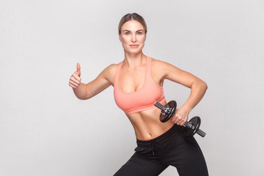 Blonde woman showing like sign and holding dumbbell. Studio shot