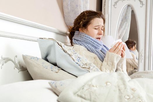 Sneezing beauty. Young sick woman with blue scarf on her neck sitting in bed and sneezing on her bed.