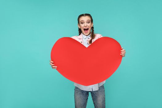 Amazed beautiful girl in casual style, pigtail hairs and jacket, standing and holding big red heart shape and looking at camera with surprised face, studio shot isolated on blue or green background