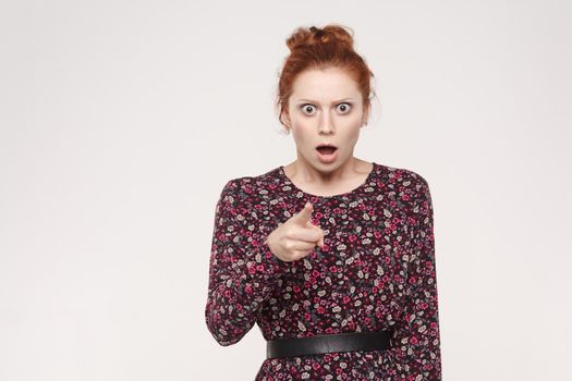 Redhead woman wearing flowers dress, opening mouths widely, having surprised shocked looks, pointing finger at camera. Isolated studio shot on gray background