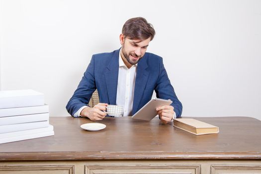 Technology, people and business concept - handsome man with beard and brown hair and blue suit and tablet pc computer and some books and cup drinking coffee or tea sitting in the office holding tablet and smile. .Isolated on white background. .