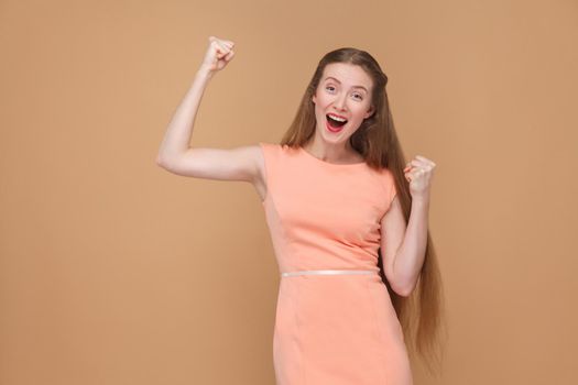 amazed winner girl showing victory sign. portrait of emotional cute, beautiful woman with makeup and long hair in pink dress. indoor, studio shot, isolated on light brown or beige background.