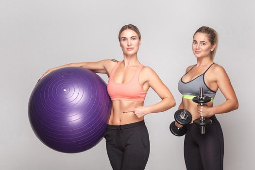 Two activity strong woman looking at camera and posing with gym equipment. Studio shot, isolated on light gray background