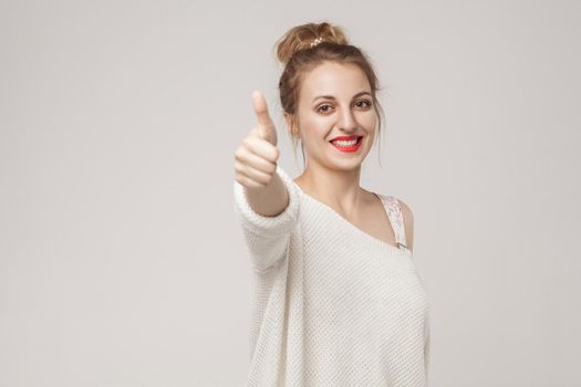 Young adult woman thumbs up, looking at camera with toothy smile. Studio shot, isolated on gray background