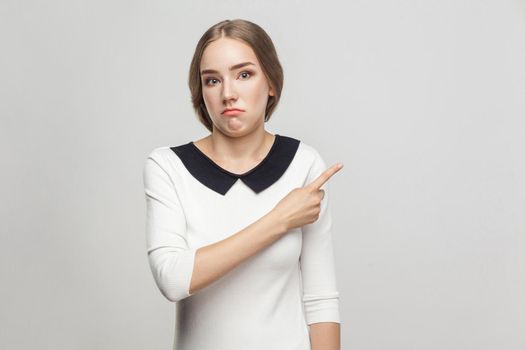 Well dressed beautiful woman with collect hair pointing finger at copy space with confused face. Studio shot, gray background
