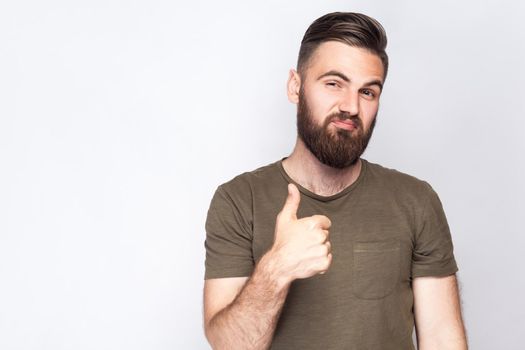 Portrait of satisfied bearded man with thumbs up and dark green t shirt against light gray background. studio shot. .