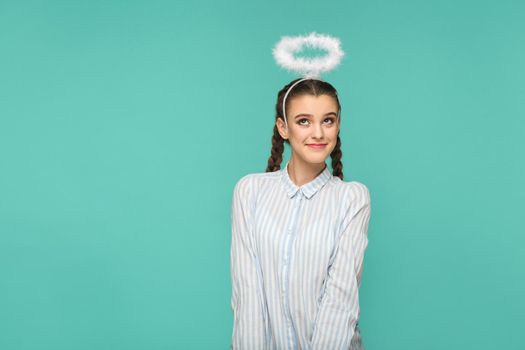 Happy funny girl in striped blue shirt and pigtail hairstyle, standing with halo on her head and looking away with smile and dreaming face, Indoor studio shot, isolated on blue or green background