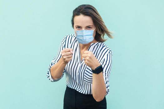 Portrait of serious middle aged woman with surgical medical mask standing with boxing fists and ready to attack . medicine and health care concept. indoor studio shot isolated on blue background.