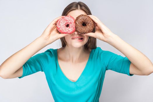 Young crazy girl bite one lips in blue blouse standing, holding, covering eyes with pink and chocolate donuts,looking through donuts like making glasses on grey background. Indoor,isolated, copy space