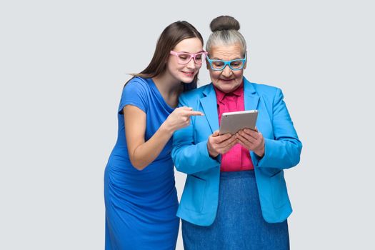 Young daughter in blue dress showing, pointing finger at tablet display in hands of aged woman. family relations between granddaughter and grandmother. indoor, studio shot, isolated on gray background