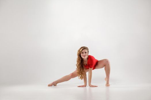 attractive athletic blonde woman with curly long hair and makeup in red leotard doing yoga pose. isolated indoor studio shot on light gray background. healthy lifestyle and leisure activity concept.