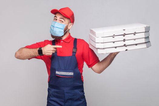 Delivery pizza on quarantine. Young man with surgical medical mask in blue uniform and red t-shirt standing, holding and pointing finger to stack of cardboard pizza boxes on grey background.