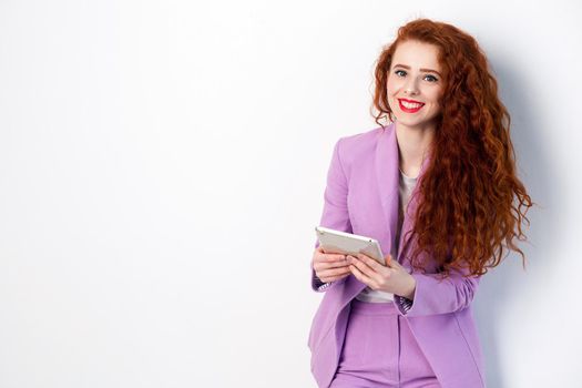 Portrait of successful happy beautiful business woman with red - brown hair and makeup in pink suit holding tablet, looking at camera with toothy smile. studio shot on gray background.