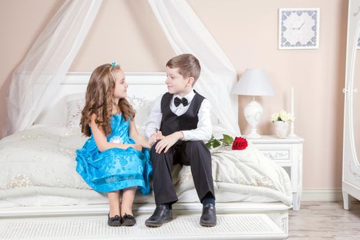 Beautiful young prince in tuxedo suit and princess holding rose in blue dress and fashion band posing sitting on the bed in bright room and looking into the distance. Love story. Studio shot.