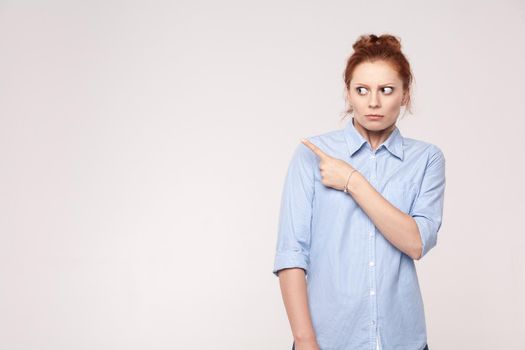 Scared redhead woman pointing away while standing isolated on gray background. Studio shot