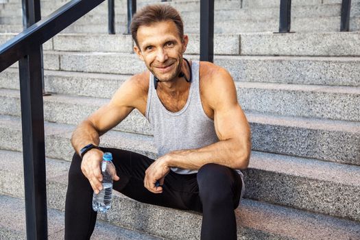 Happy smily sportsman resting and holding bottle of water, sitting on steps looking at camera and smiling . fitness, sport, exercising and people healthy lifestyle concept.
