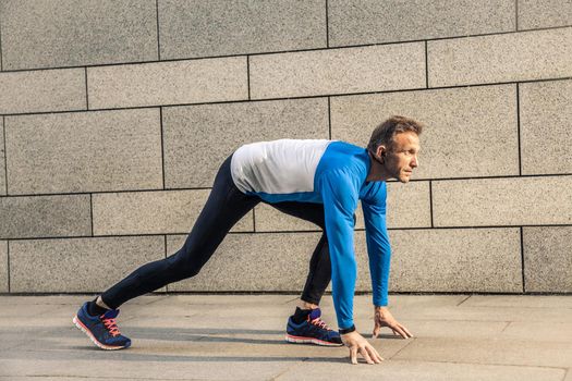 Handsome middle aged serious man in blue black sports uniform and headphones and fitness tracker is running in the city.