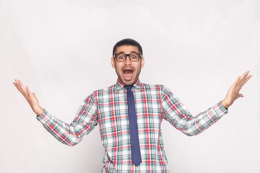 Amazed or shocked bearded businessman in checkered shirt, blue tie and eyeglasses standing, raised hand and looking at camera with surprised open mouth. studio shot, isolated on light grey background.