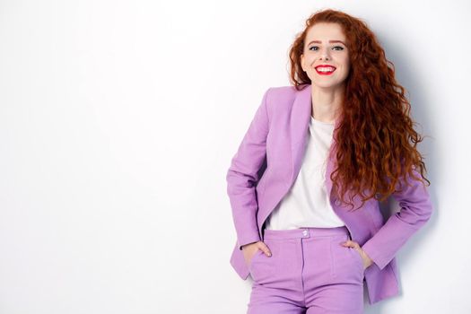 Portrait of successful happy beautiful business woman with red - brown hair and makeup in pink suit. looking at camera with toothy smile, studio shot on gray background.
