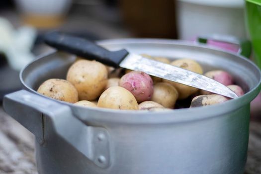 Stainless steel saucepan with raw new potatoes and a kitchen knife. Potatoes not peeled. Close-up of yellow and red skinned potatoes. Peeling potatoes in the kitchen on a wooden rustic table