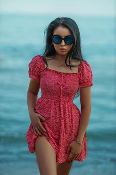 Tanned sexy brunette model in a red dress with small polka dots and round big glasses for sun protection posing on the seashore