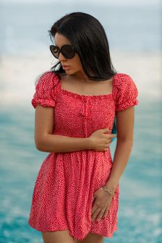 Tanned sexy brunette model in a red dress with small polka dots and round big glasses for sun protection posing on the seashore