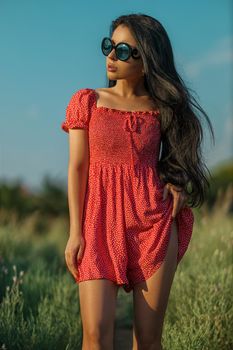 Tanned sexy brunette girl in a red dress with small polka dots posing barefoot in a meadow among wild herbs, flowers and trees on a summer sunny day