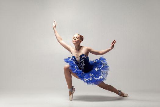 Young beautiful ballerina with bun collected hair wearing blue dress and pointe shoes dancing gracefully isolated on white background. indoor, studio shot.