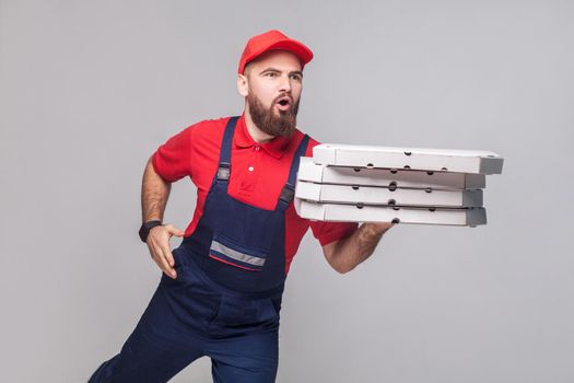 Hurry up! Young amazed delivery man with beard in blue uniform and red t-shirt holding stack pizza boxes and running to delivery on grey background. Indoor, studio shot, isolated, copy space.