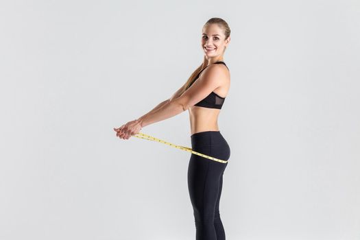 Weight loss concept. Happiness blonde woman holding centimeter on her ass. Indoor shot, gray background