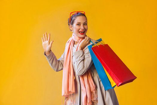 Hello! Happiness young adult woman with freckles, collect hair, looking at camera, hand up , holding many shopping bag, enjoying good shopping day. Studio shot, yellow background