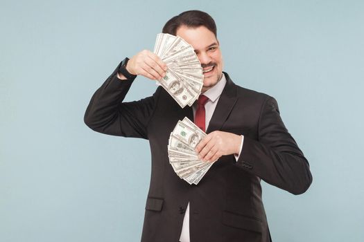 Businessman hides behind fans out of money. Indoor, studio shot, isolated on light blue or gray background