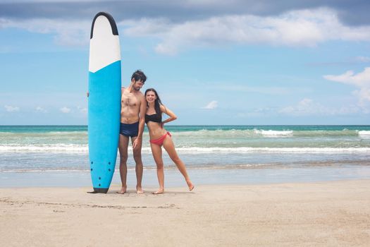 Happy young couple standing with a surfboard on the beach. Stock image.