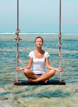 The girl meditates on a swing in the ocean beach in Bali, Indonesia. Stock image.