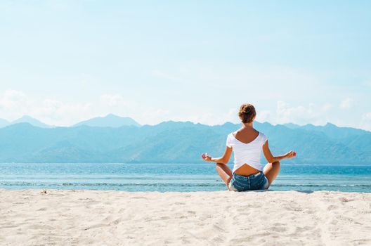 A young woman meditates in a pose of yoga on a sandy beach overlooking the blue ocean and the mountains. Stock image.