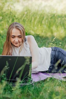 On the banner, a young girl works with a laptop in the fresh air in the park, sitting on the lawn. The concept of remote work. Work as a freelancer. The girl takes courses on a laptop and smiles