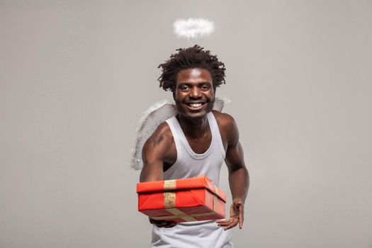 Love concept and sharing goft. dark skinned bearded African angel funny man with white shirt, nimbus and winks atanding and holding present gift box. its for you. Indoor, isolated on gray background