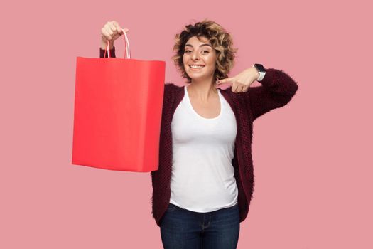 Cheerful beautiful young woman in blue shirt with curlty hairstyle standing and pointing finger to shopping bags and toothy smile, looking at camera. Studio shot, pink background, isolated, indoor