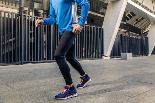 Handsome middle aged serious man in blue black sports uniform and headphones and fitness tracker is running in the city.