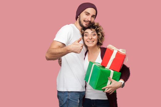 Portrait of happy couple of friends in casual style standing, hugging, hold two boxing present, showing thumb up and toothy smile, celebrate anniversary. Isolated, indoor, studio shot, pink background