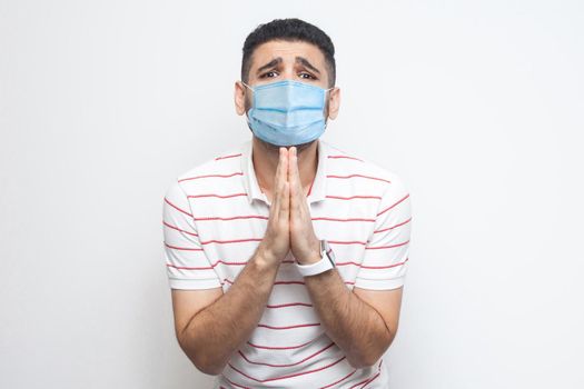 Please help me. Portrait of sad alone man with surgical medical mask in striped t-shirt standing, palm hands and looking at camera pleading or begging. indoor studio shot, isolated on white background
