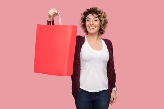 Satisfied modern young woman in blue shirt with curlty hairstyle standing and showing shopping bags and toothy smile, looking at camera. Studio shot, pink background, isolated, indoor, copy space