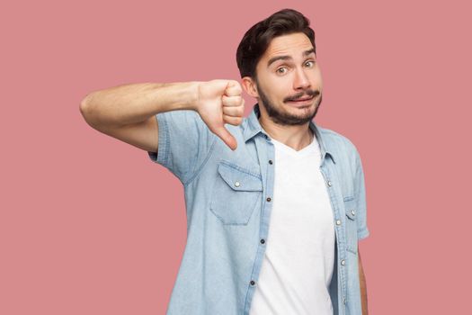No, Dislike. Portrait of dissatisfied handsome bearded young man in blue casual style shirt standing, thumbs down and looking at camera . indoor studio shot, isolated on pink background.