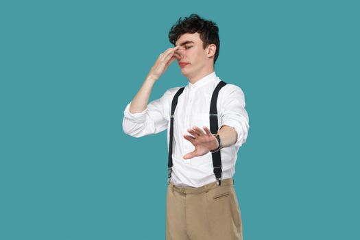 Confused man showing stop and pinching his nose. Portrait of handsome hipster curly young businessman in classic casual white shirt, suspender standing. indoor studio shot isolated on blue background.