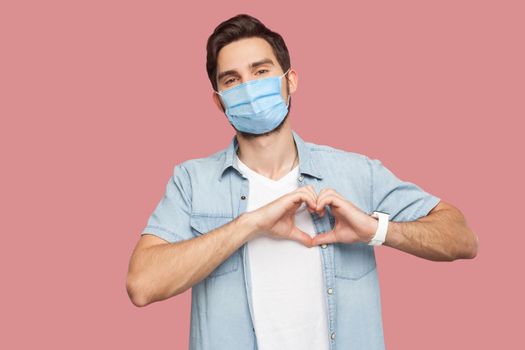 Portrait of happy smiley young man with surgical medical mask in blue shirt standing with love hand gesture on heart and looking at camera smiling. indoor studio shot, isolated on pink background.