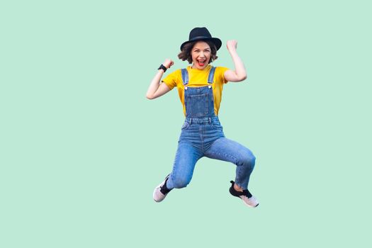 Portrait of screaming happy suprised young girl in blue denim overalls, yellow shirt and black hat jumping, looking and celebraiting her victory. indoor studio shot isolated on light green background.