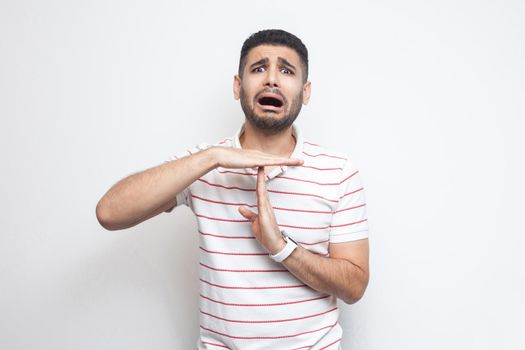 I need more time. Portrait of pleased handsome bearded young man in striped t-shirt standing with time out gesture and looking at camera pleading. indoor studio shot, isolated on white background.