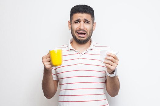 Unhappy worry sick young adult man in white t-shirt standing and holding yellow cup with white napkin, drink hot tea, has flu. Indoor, isolated, white background, studio shot