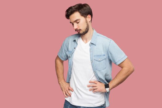 Portrait of sad, unhappy or upset handsome bearded young man in blue casual style shirt standing with hands on waist, holding his head down, depressed. indoor studio shot, isolated on pink background.